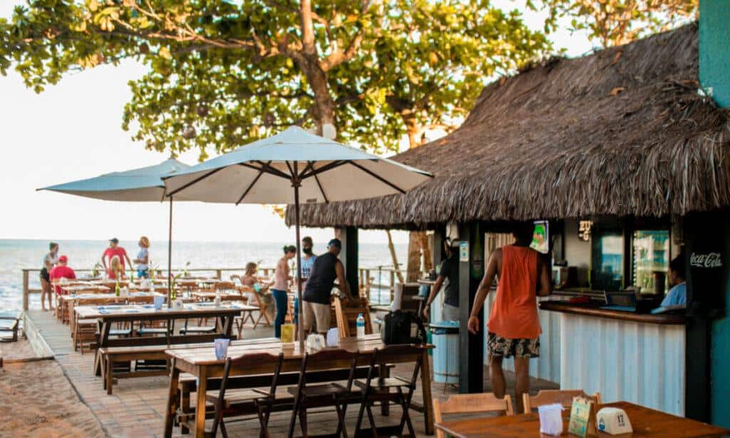 Casal de mesas ao ar livre em restaurante próximo à praia com vista para o mar e sombra de guarda-sol, ambiente aconchegante e casual na Casa Maria Maria.