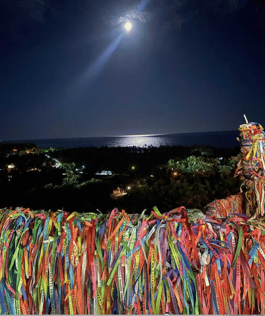 Cordão de fitas coloridas na varanda de Casa Maria Maria com vista para o mar sob a luz da lua, criando um ambiente acolhedor, religioso e cultural, perfeito para momentos de reflexão e celebração na natureza.