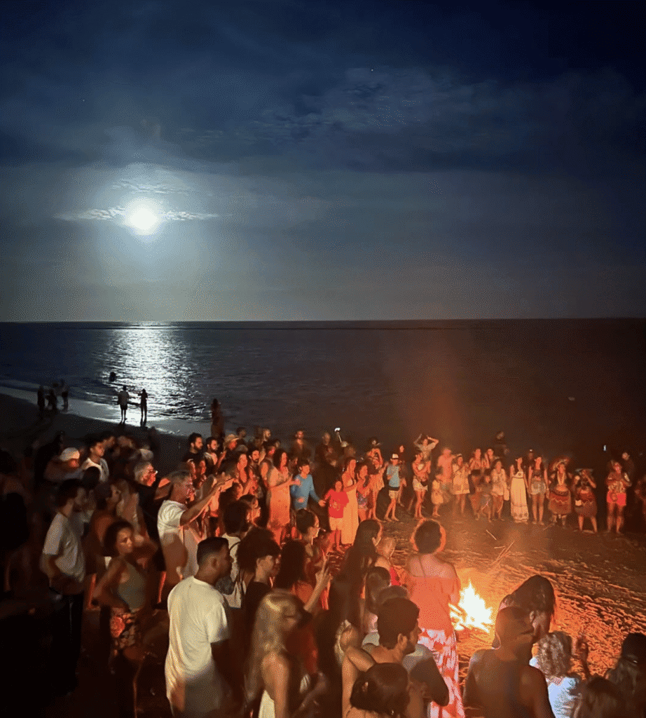 Alto astral de uma festa na praia à noite sob a luz do luar, com diversas pessoas ao redor de uma fogueira, transmitindo alegria, confraternização e clima de lazer na costa brasileira.