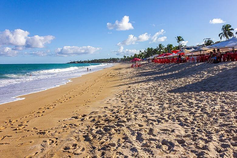 Cadeira de praia na praia de areia dourada com guarda-sóis vermelhos, palmeiras e céu azul claro, ideal para férias e lazer na ilha, perto da Casa Maria Maria no Maranhão, Brasil.